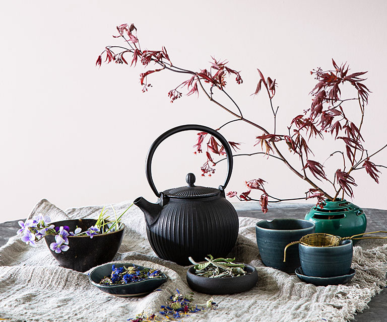 A charcoal-gray cast iron tea pot, bowls, and a flowering tree stem against a gray-violet-painted wall.