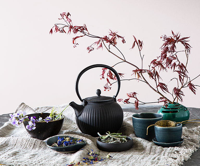 Une théière en fonte gris anthracite, des bols et une tige d’arbre en fleurs contre un mur peinturé en violet-gris.