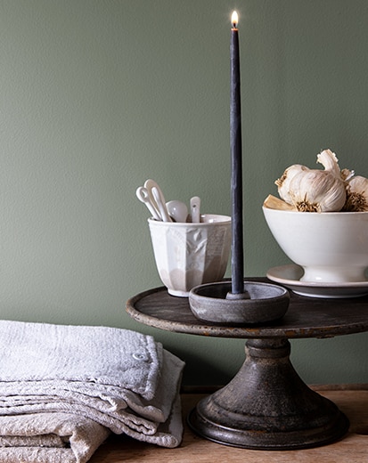 Contre un mur peint d’un vert riche, un plat à gâteau présente une bougie effilée allumée à côté de deux bols en porcelaine.