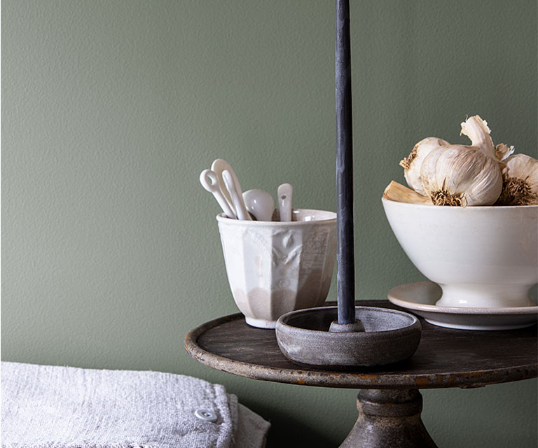 Against a rich green-painted wall, a cake plate holds a lit tapered candle beside two porcelain bowls.