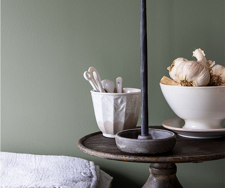 Contre un mur peint d’un vert riche, un plat à gâteau présente une bougie effilée allumée à côté de deux bols en porcelaine.