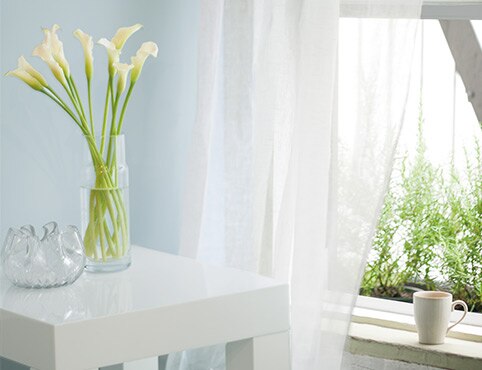 A vase of flowers sits on a small, square bedside table next to an open window framed by a gauzy white curtain and a light blue wall.