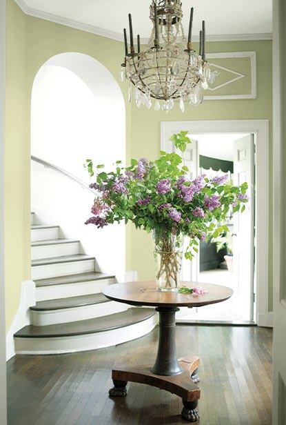 Warm green entryway with chandelier above wood circle table with a bouquet of flowers.