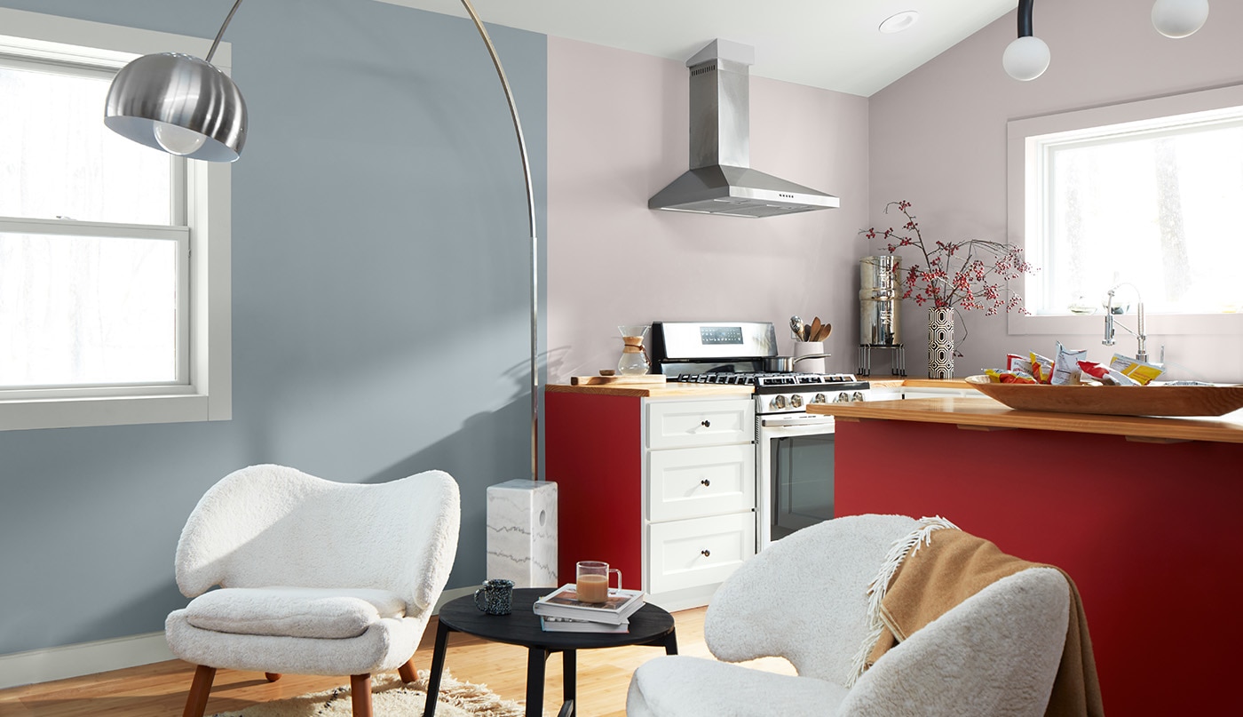 Open living room and kitchen with light blue and lavender-gray walls, a white ceiling, a set of modern white armchairs, and cabinets painted red and white.