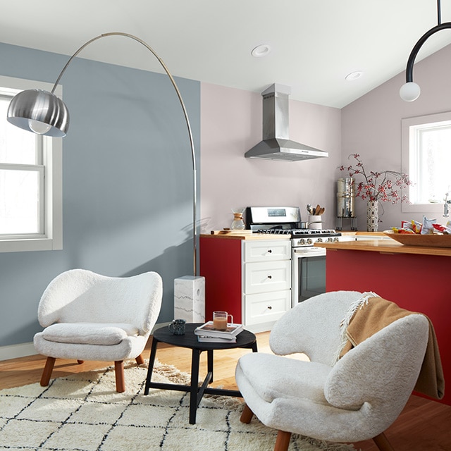 Open living room and kitchen with light blue and lavender-gray walls, a white ceiling, a set of modern white armchairs, and cabinets painted red and white.