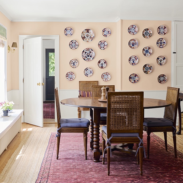 Dining room with pale pink on the upper portion of the walls and white on the lower portion; a wooden bench is set beneath large, bright windows that illuminate a wooden dining table set on a red rug and light wood flooring.