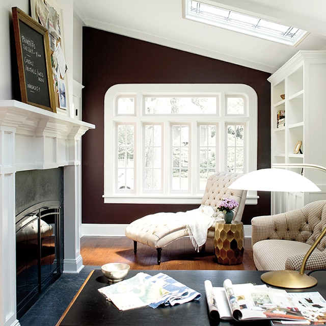 A study with white walls and a chocolate brown accent wall set with a large, white-framed window, a fireplace, a desk and several other pieces of furniture spread throughout the room.