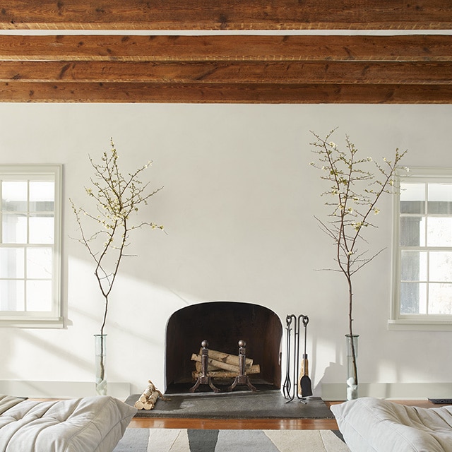 Living room with fireplace centred on a white wall, framed by two matching windows, two plants, and two modern white chairs, with brown wood joists along the ceiling.