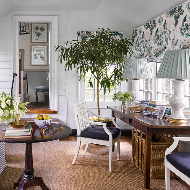 Une véranda aux murs en planches à feuillure peintes en blanc avec de grandes fenêtres où sont accrochés des rideaux fleuris éclaire un assortiment de meubles et de plantes en pot. 