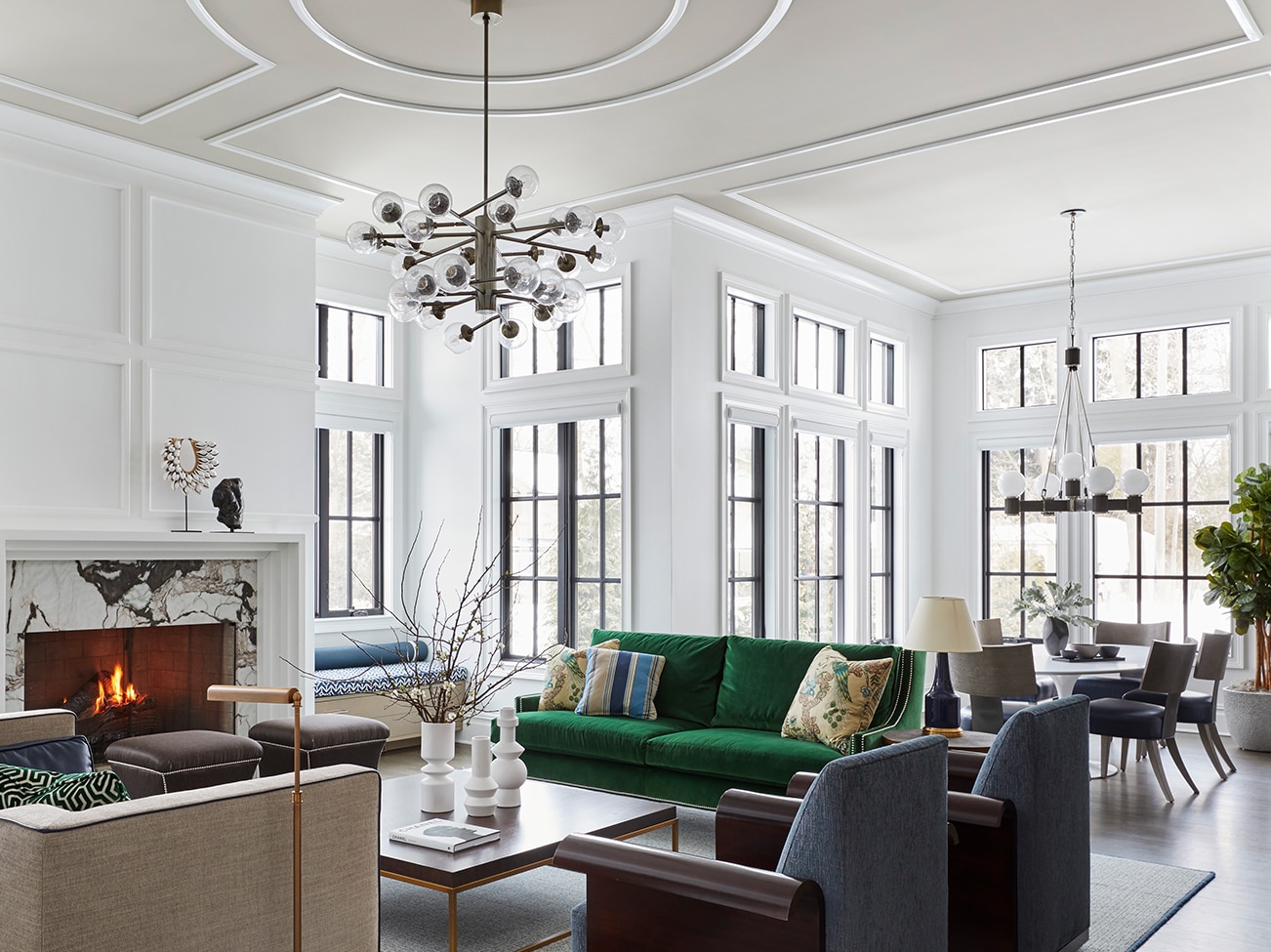 Open living room in white with marble fireplace, taupe ceiling, green sofa, and beige and gray seating next to a sleek dining area.