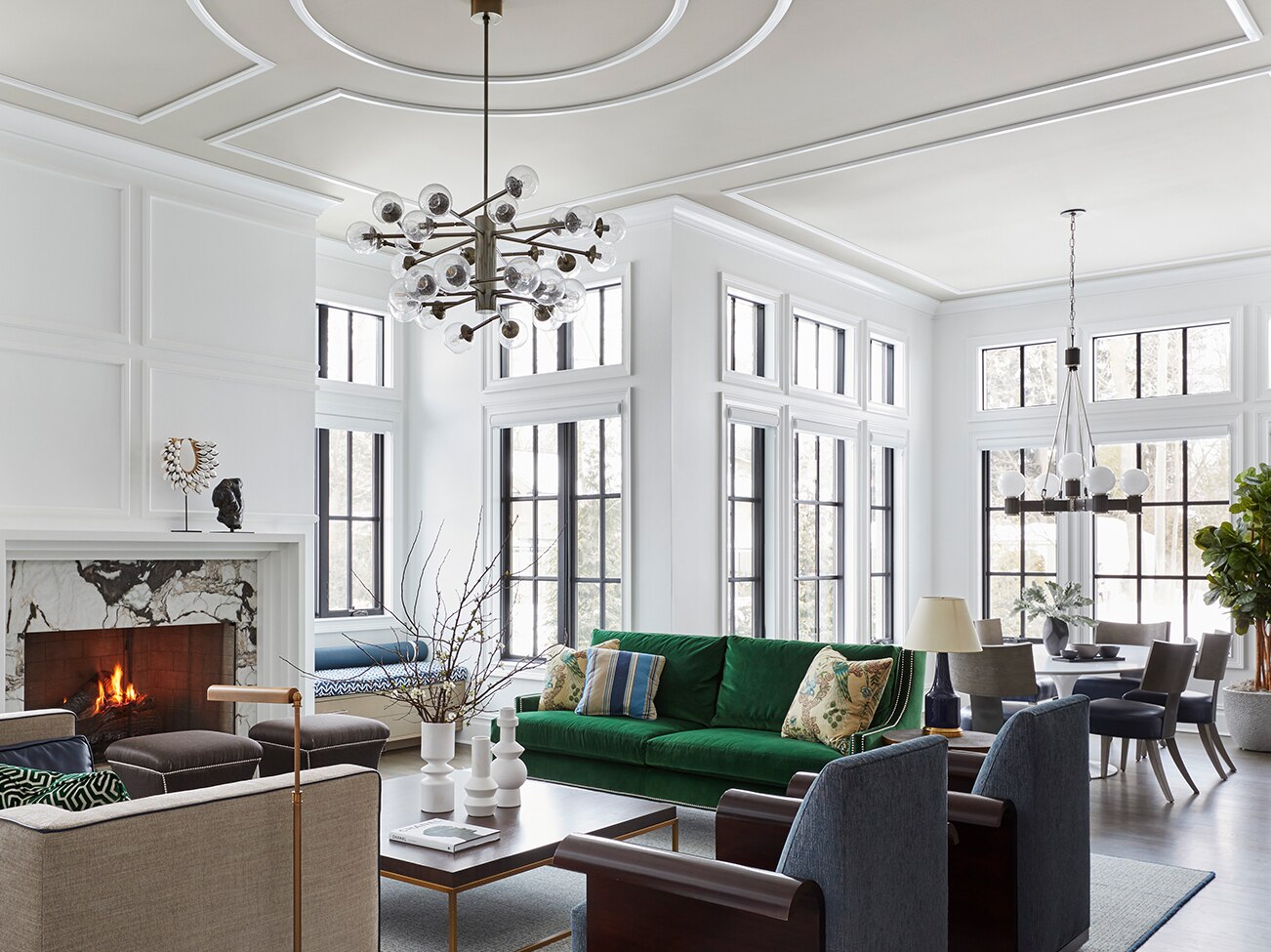 Salon blanc à aire ouverte avec cheminée en marbre, plafond de couleur taupe, sofa vert et fauteuils beiges et gris qui le séparent de l’élégante salle à manger.