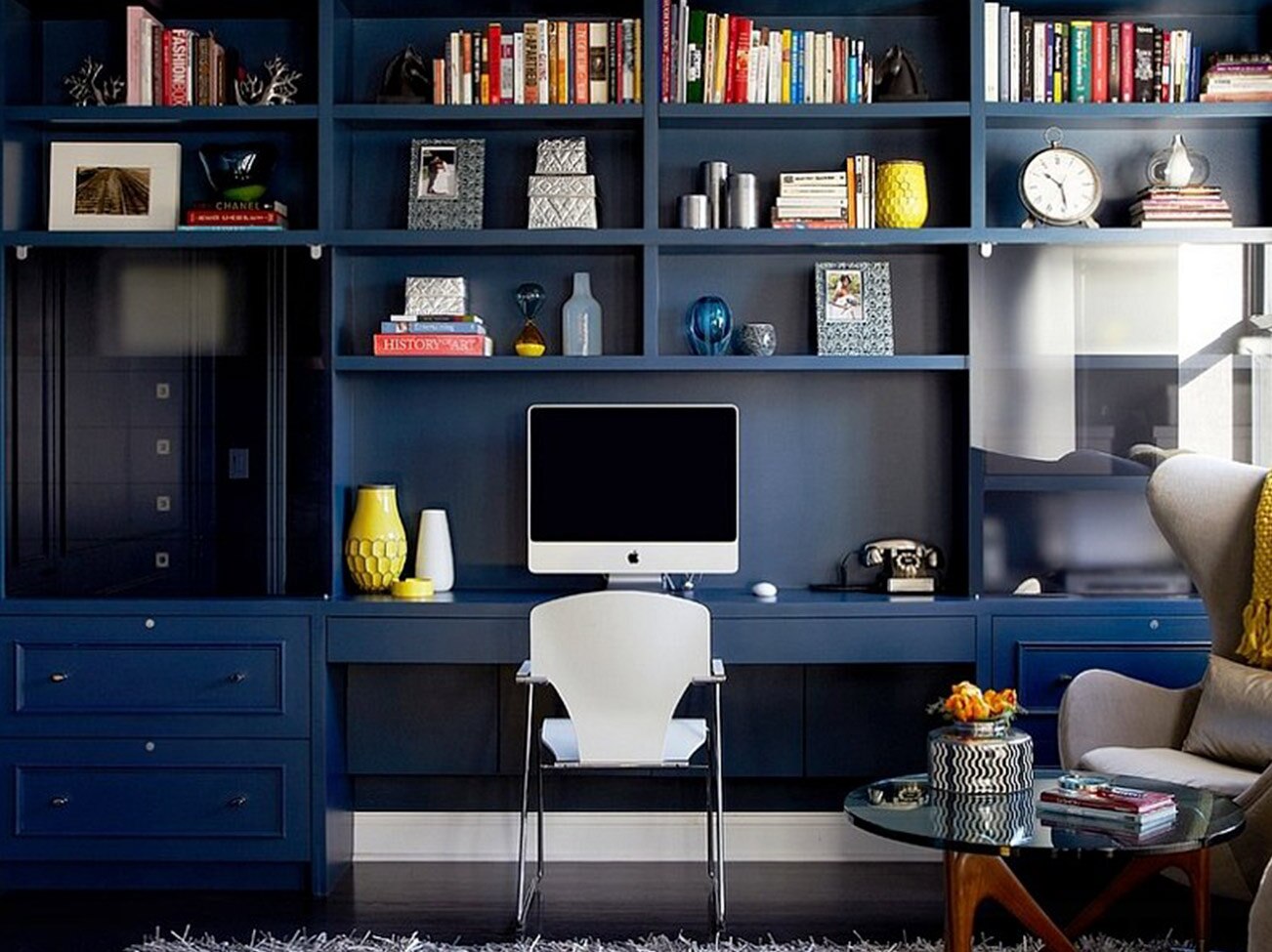 Bureau à domicile avec étagères et table encastrées bleu foncé, coin-détente aux fauteuils modernes et petite table décorative en verre.