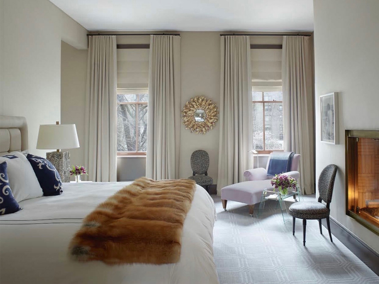 Élégante chambre à coucher blanche avec foyer allumé, lit avec jeté en fourrure, coussins décoratifs bleus et chaise longue.