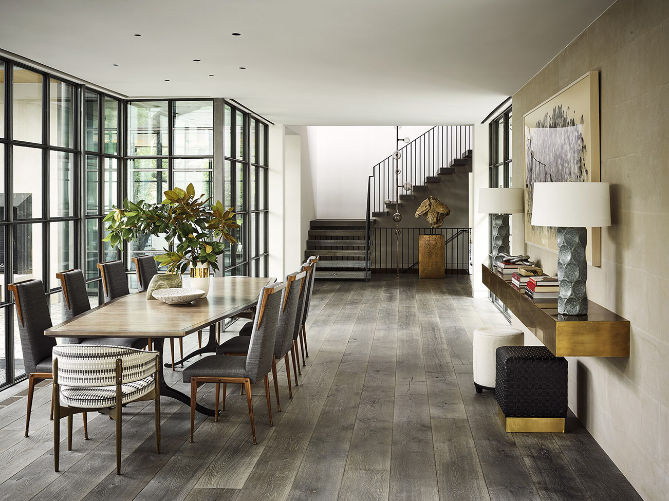 Dining room with grey wood floors, floor-to-ceiling metal-paned windows, and elegant staircase.