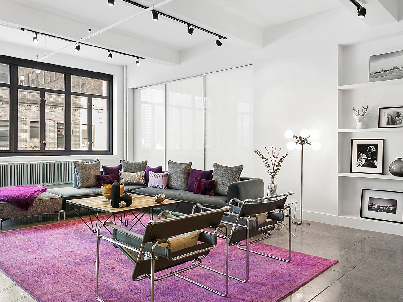 Living room, metal-framed windows, black track lighting, gray sectional, Breuer chairs and fuchsia area rug on stone floor.