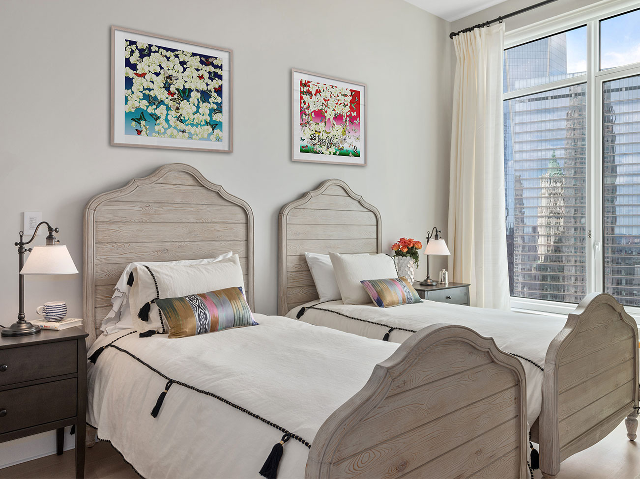 Bedroom with twin beds, artwork over headboards, pale gray walls, city view through sheer shades and white curtains.