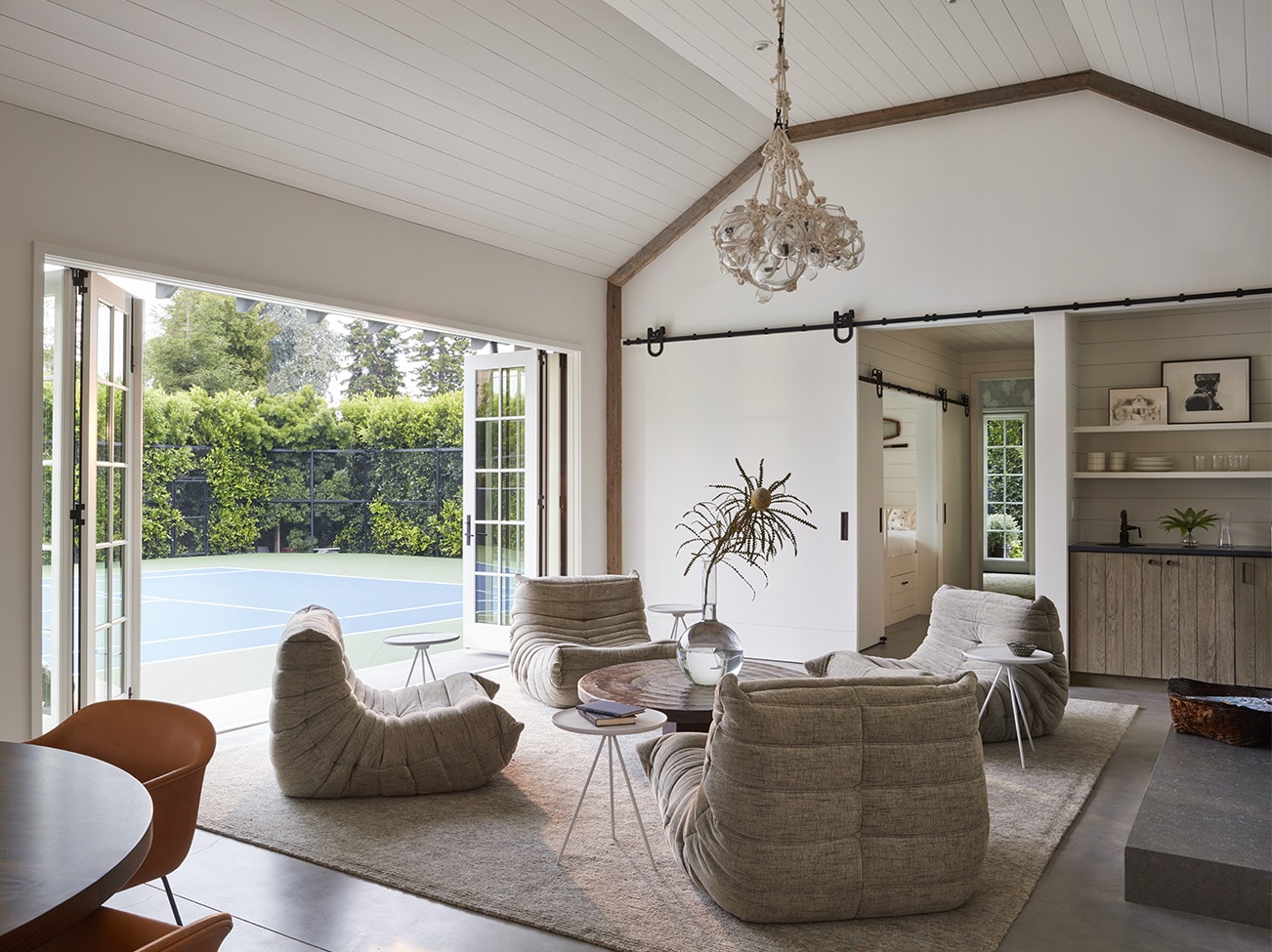 Coin-détente dans une pièce de style grange avec quatre fauteuils, un plafond de bois en mansarde, un luminaire à suspension et des portes-fenêtres qui ouvrent sur un court de tennis.