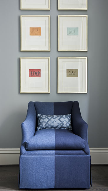 Sitting area with pale blue wall with six white framed prints above a two-toned blue arm chair with a blue batik pillow.