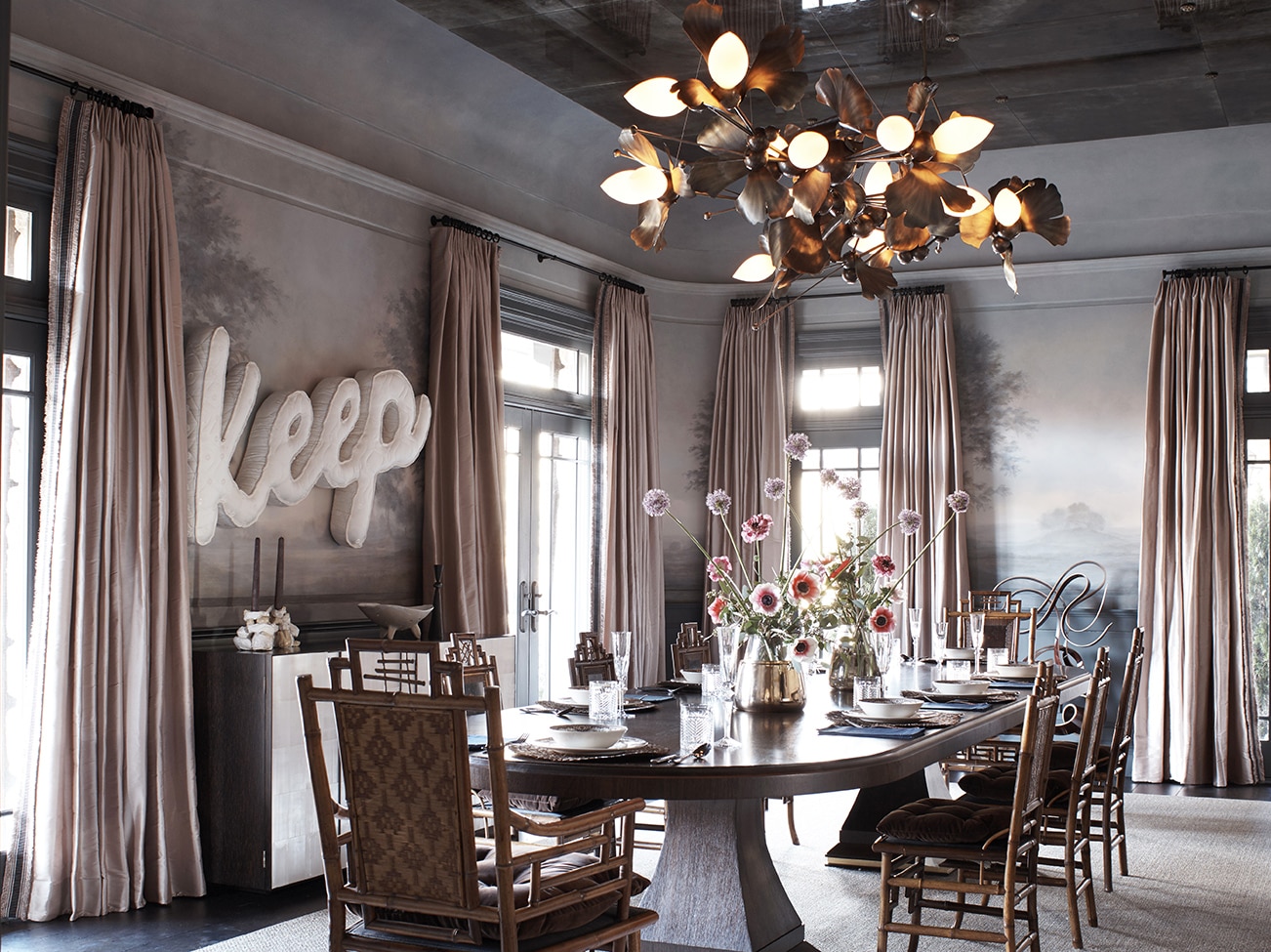 Salle à manger avec grandes fenêtres, rideaux pâles, papier peint et décorations sur les murs, plafond en miroir, lustre en forme de fleurs, table et chaises en bois.