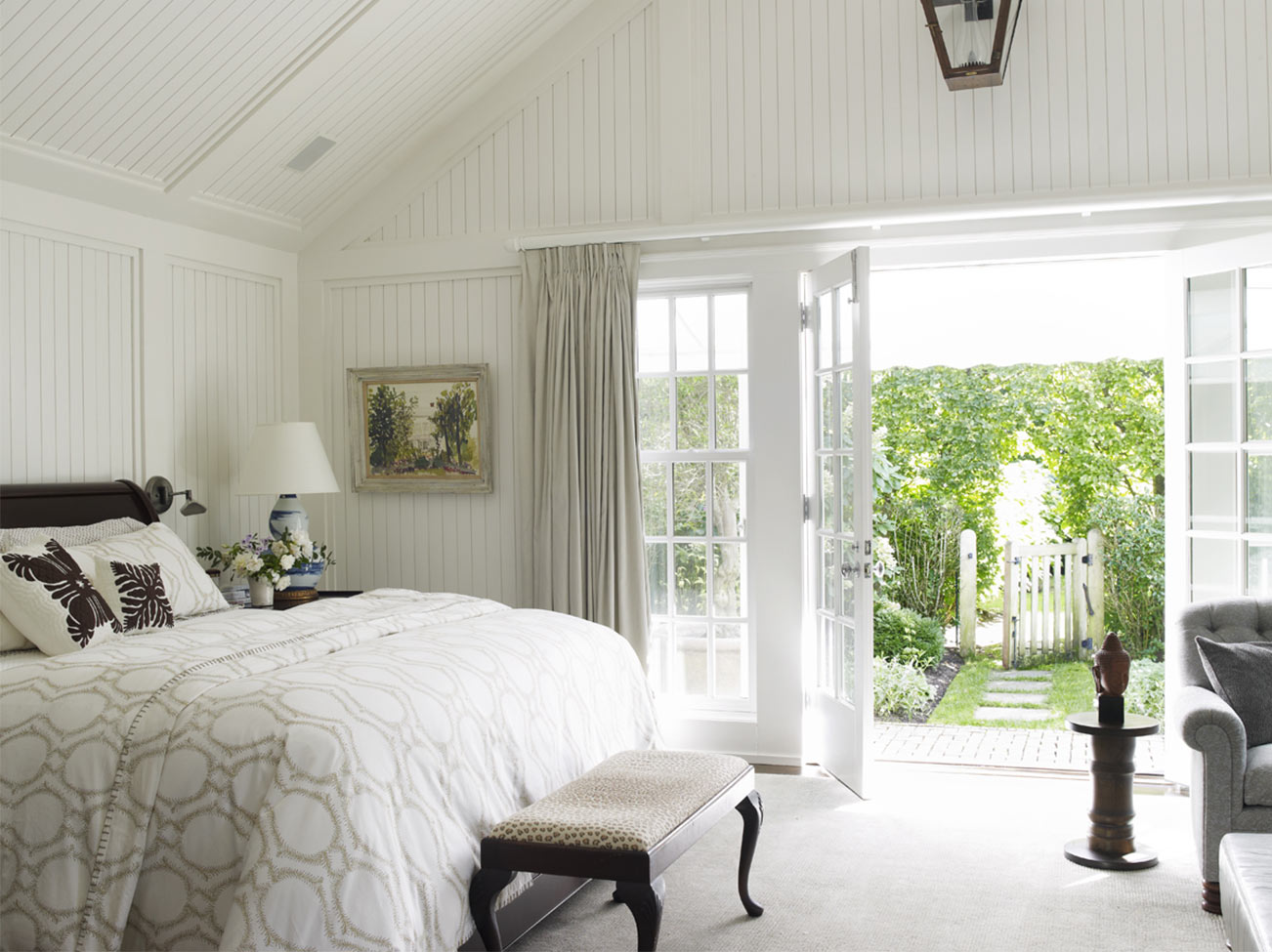 Chambre à coucher en planches à baguette blanches, literie raffinée et banc au pied du lit, fauteuil gris et portes-fenêtres qui ouvrent sur un jardin.
