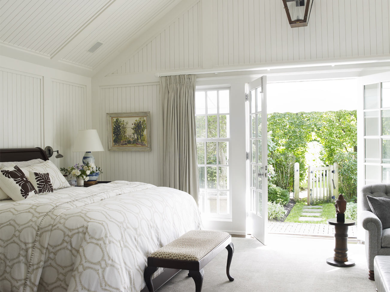 Bedroom with white beadboard throughout, elegant bedding and bedroom bench, gray arm chair and French doors open to a garden. Bedroom with white beadboard throughout, elegant bedding and bedroom bench, gray arm chair and French doors open to a garden.