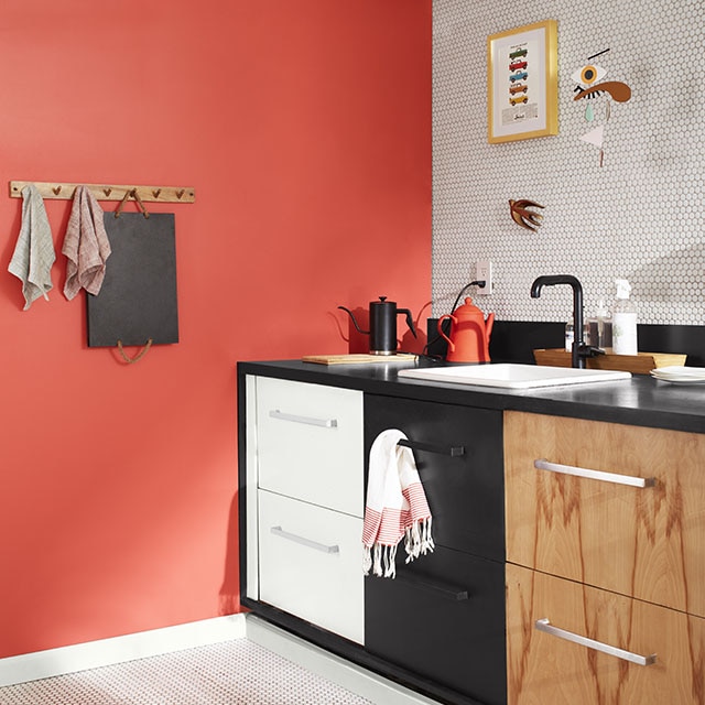 Un elegante rincón de la cocina con una pared pintada de coral, el piso y una pared antisalpicaduras con azulejos blancos, una pared superior blanca, tuberías a la vista y tres conjuntos de cajones: uno pintado de blanco, el segundo de negro y el tercero de madera.