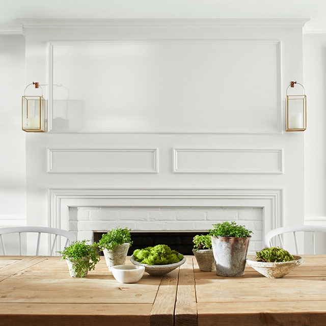 An elegant white dining room with white-painted brick fireplace with lanterns above mantel, and wooden table with several potted succulents on top of it.