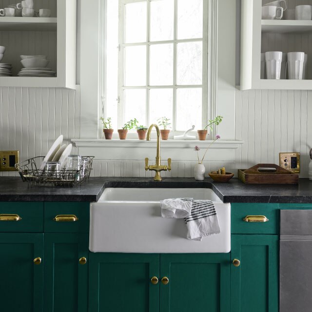 A country kitchen with white-painted walls and forest green-painted lower cabinets under a farmhouse sink beneath a large paned window. 