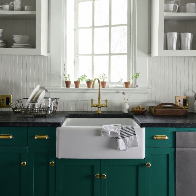 A country kitchen with white-painted walls and forest green-painted lower cabinets under a farmhouse sink beneath a large paned window.  