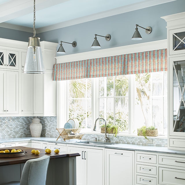 Una cocina acogedora con paredes pintadas de azul claro y techo pintado de un azul glacial que enmarca vigas blancas, tres ventanas grandes sobre un lavabo, una isla de cocina y gabinetes pintados de blanco.