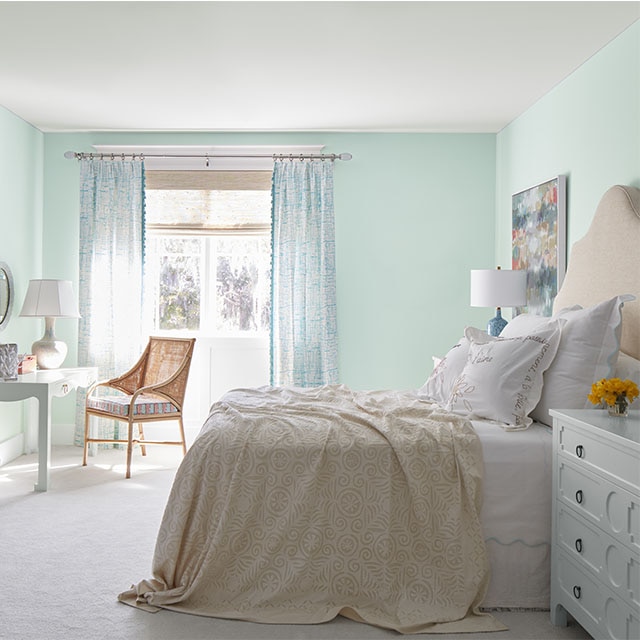 A beachy bedroom painted in a minty green hue with white ceiling, white-painted dresser, and off-white carpeting. 