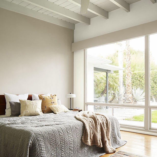 A bedroom painted in neutral hues and white-painted shiplap ceiling with exposed beams, ceiling fan, and a floor-to-ceiling window wall with views of desert landscaping.