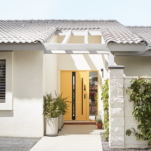 A sunny home exterior painted in off-white with  a yellow front door, cacti and other dessert landscaping.