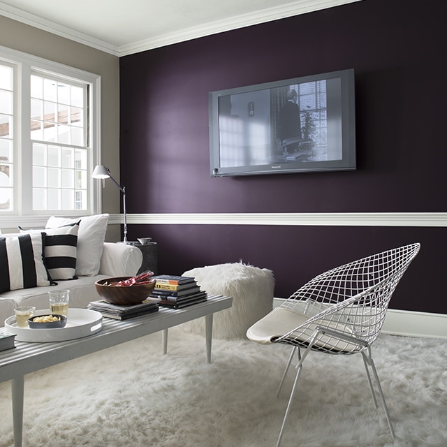 A contemporary living room with a dark purple accent wall frames a flat-screen TV, a white couch with black and white throw pillows, white metal chair, shaggy white rug, and two faux fur ottomans.
