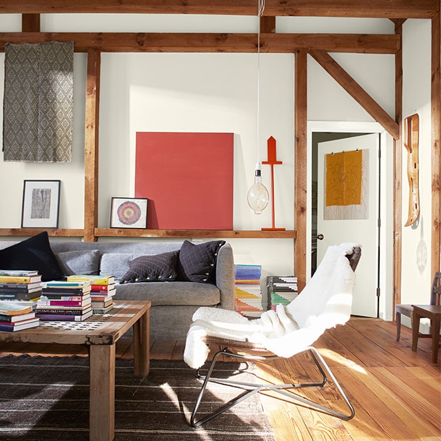 Exterior light streams into an inviting living room painted in white hues, exposed wood beams, gray couch, wood floor, and pops of color throughout.