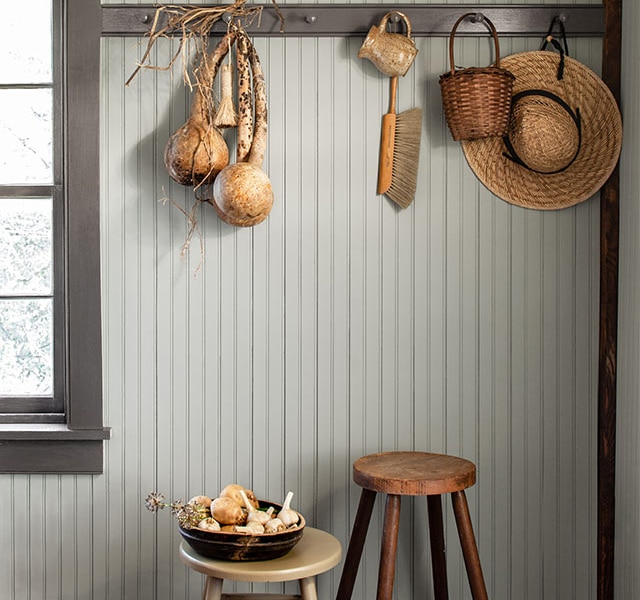 A gray-painted beadboard wall with brown trim and wall pegs holding a straw hat, a basket, brush, and other rustic trinkets hanging over two wooden stools.