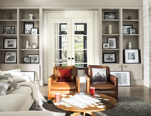 A comfy living room with gray-painted  built-in bookshelves, white french doors, a white sofa, and brown leather chairs.