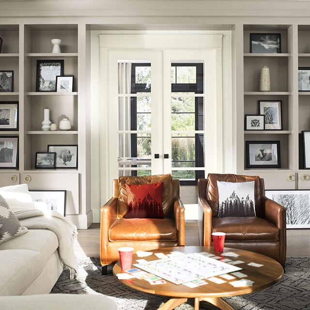 A comfy living room with gray-painted  built-in bookshelves, white french doors, a white sofa, and brown leather chairs.