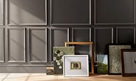 Artwork and frames arranged on a floor against a deep chocolate brown-painted wall with decorative molding.