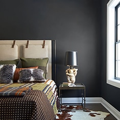 Black bedroom walls with a white and brown cow print rug underneath a tan bed with a printed comforter.