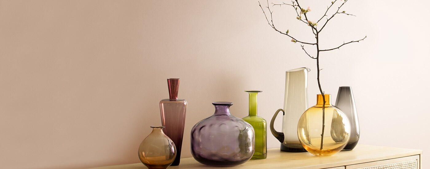 Dusty pink walls behind a rattan cabinet with an array of glass vases placed on top.