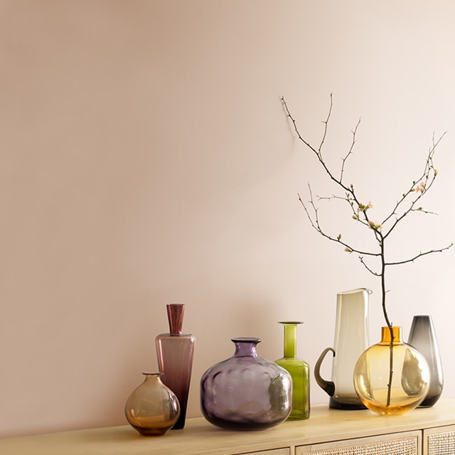 Dusty pink walls behind a rattan cabinet with an array of glass vases placed on top.