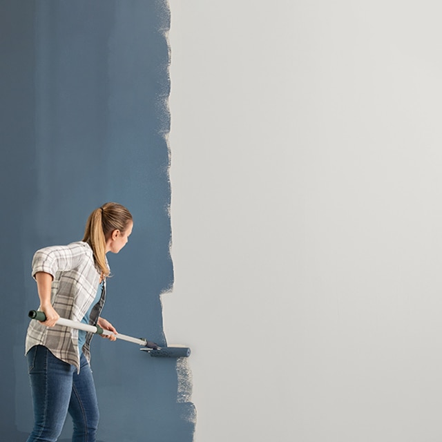 A woman rolls blue Benjamin Moore paint onto a white wall.