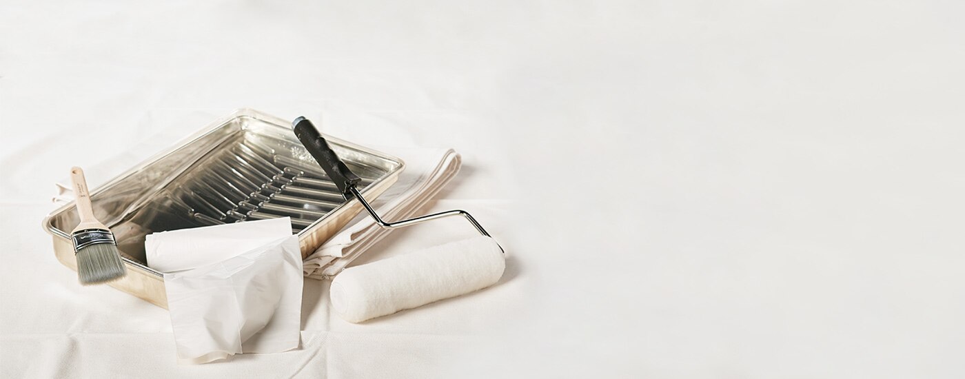 Painting supplies including a metal paint tray, an angled sash brush, a paint roller and a plastic drop cloth.