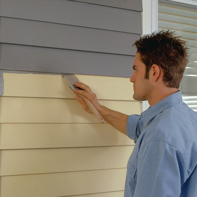 Homme appliquant de la peinture grise sur le parement en vinyle d’une demeure.