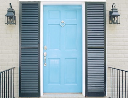 A painted front door in Bahamian Sea Blue framed by dark gray shutter on an off-white brick home facade.