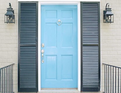 Una puerta principal pintada de color Azul Mar de Bahamas enmarcada por contraventanas de color gris oscuro sobre una fachada de ladrillos de color blanco impuro.