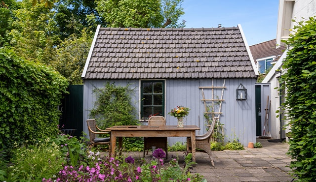 A romantic blue shed next to a long wooden dining table, three rattan chairs, a pitcher with flowers, a trellis, and plants.