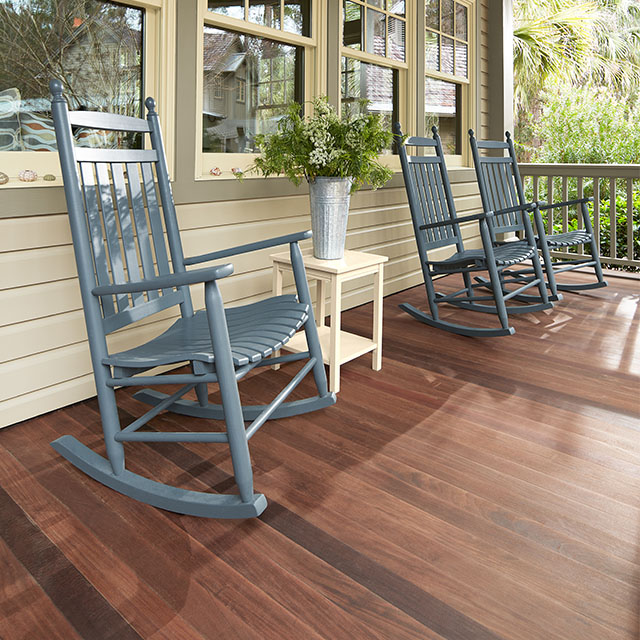 Blue rocking chairs show off nicely on this covered porch with a stained wood floor, greige siding and multiple muted-green trimmed windows.