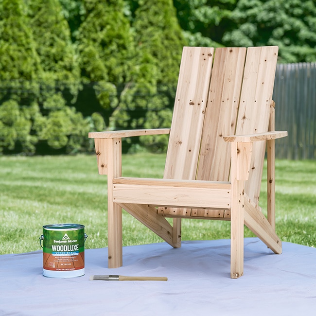 A 3.79 L can of Woodluxe® Exterior Stain and a paintbrush next to a wooden Adirondack chair.